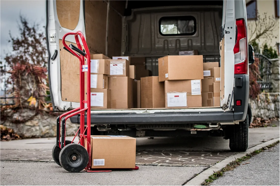 Beladener Transporter mit offenen Hecktüren, davor eine Sackkarre mit Karton
